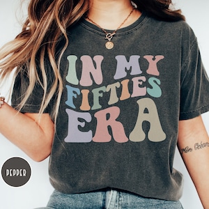 May include: A dark gray t-shirt with the text "In My Fifties Era" in colorful retro lettering. The shirt is made of a soft, comfortable material and has a relaxed fit.