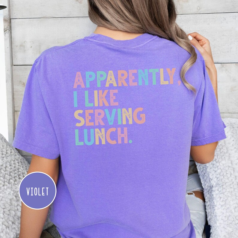 May include: A violet t-shirt with the text "APPARENTLY, I LIKE SERVING LUNCH" in pastel colors. The shirt is a solid violet color and has a crew neck. The text is in a playful font.