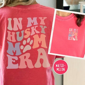 May include: A pink t-shirt with the text "In My Husky Mom Era" in a retro font with paw prints. The text is in shades of pink, purple, and gray.