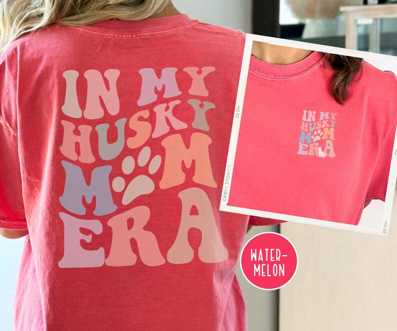 May include: A pink t-shirt with the text "In My Husky Mom Era" in a retro font with paw prints. The text is in shades of pink, purple, and gray.