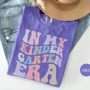 May include: Purple short-sleeved t-shirt with the text "In My Kindergarten Era" in rainbow colors. The text is in a retro font style.