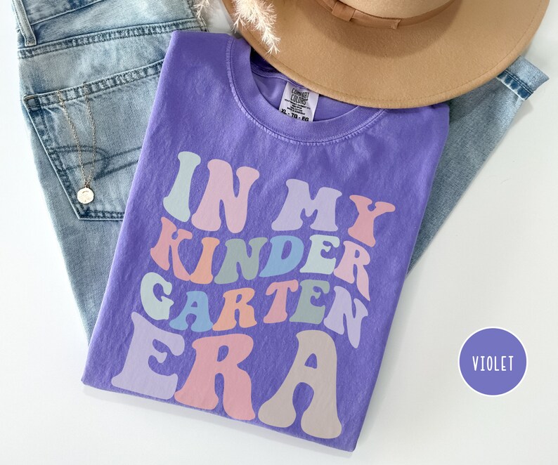 May include: Purple short-sleeved t-shirt with the text "In My Kindergarten Era" in rainbow colors. The text is in a retro font style.
