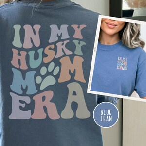 May include: A blue jean t-shirt with the text "In My Husky Mom Era" in colorful retro letters. The shirt is shown on a model wearing the shirt.