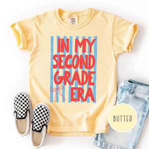 May include: A pale yellow short-sleeve t-shirt featuring a graphic with vertical light blue stripes and the red text "IN MY SECOND GRADE ERA" with a small red bow illustration. The shirt is displayed alongside black and white checkered slip-on shoes.