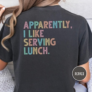 May include: Dark gray t-shirt with the text "Apparently, I Like Serving Lunch" in pastel colors. The shirt is a casual style, suitable for everyday wear.