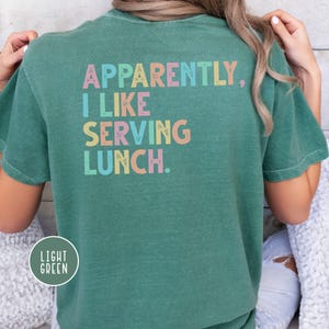 May include: Light green t-shirt with the text "Apparently, I Like Serving Lunch" in pastel colors. The shirt has a crew neck and short sleeves. The words are in a playful, colorful font.