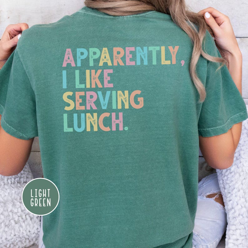 May include: Light green t-shirt with the text "Apparently, I Like Serving Lunch" in pastel colors. The shirt has a crew neck and short sleeves. The words are in a playful, colorful font.