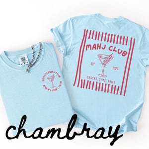 Puede incluir: Dos camisetas azul claro con gr&aacute;ficos rojos. Una camiseta tiene un logotipo frontal con una copa de c&oacute;ctel y el texto "EMILY'S MAHJ CLUB". La otra camiseta tiene un gr&aacute;fico trasero con una copa de c&oacute;ctel y el texto "MAHJ CLUB EST 2026 CRACKS, DOTS, BAMS".