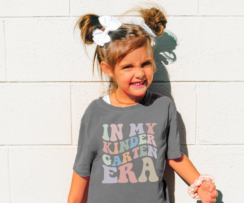 May include: A child wearing a gray t-shirt with the text "In My Kinder Garden Era" in rainbow colors. The child has their hair in two pigtails with white bows.
