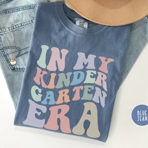 May include: A blue jean t-shirt with the text "In My Kindergarten Era" in rainbow colors. The text is in a retro font style.
