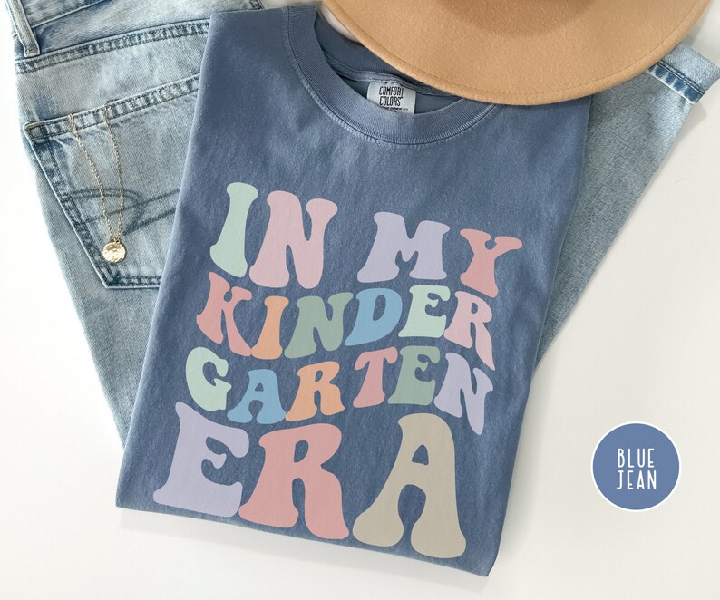 May include: A blue jean t-shirt with the text "In My Kindergarten Era" in rainbow colors. The text is in a retro font style.
