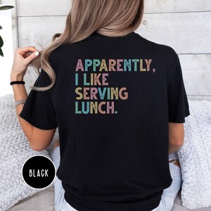May include: Black t-shirt with the text "APPARENTLY, I LIKE SERVING LUNCH" in pastel colors. The shirt has a crew neck and short sleeves. A black circle with the word "BLACK" is at the bottom left.