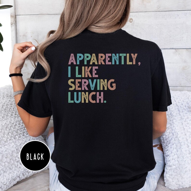 May include: Black t-shirt with the text "APPARENTLY, I LIKE SERVING LUNCH" in pastel colors. The shirt has a crew neck and short sleeves. A black circle with the word "BLACK" is at the bottom left.