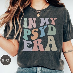 May include: A dark gray t-shirt with the text "IN MY PSY ERA" in colorful, retro-style lettering. The shirt is worn by a person with long brown hair and a gold necklace.