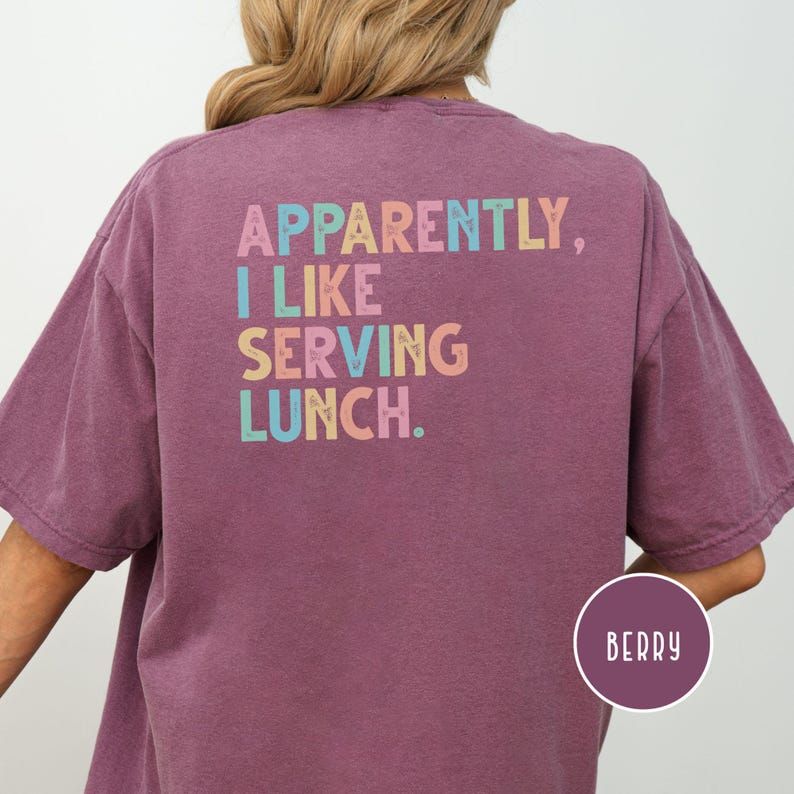 May include: Berry-colored t-shirt with the text "Apparently, I like serving lunch" in colorful letters. The shirt has short sleeves and a relaxed fit. A circular label at the bottom right corner reads "Berry".