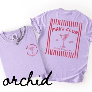 Puede incluir: Dos camisetas color orqu&iacute;dea con gr&aacute;ficos rojos. Una camiseta presenta un peque&ntilde;o dise&ntilde;o circular con una copa de c&oacute;ctel y texto. La otra camiseta tiene un dise&ntilde;o m&aacute;s grande en la espalda con una copa de c&oacute;ctel y el texto "MAHJ CLUB".