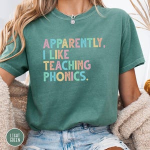 May include: A light green t-shirt with the text "Apparently, I Like Teaching Phonics" in pastel colors. The shirt is a crew neck style and has short sleeves. The words are in a playful font.