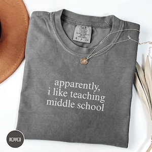 Puede incluir: Una camiseta gris carbón de manga corta con cuello redondo y una etiqueta de la marca Comfort Colors. La prenda presenta el texto impreso en blanco "apparently, i like teaching middle school" centrado en el pecho. Incluye un collar de tono dorado.