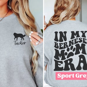 May include: A grey sweatshirt with a black silhouette of a Bernese Mountain Dog and the name "Tucker" on the front. The back of the sweatshirt has the text "In My Bernese Mom Era" in a retro font. The sweatshirt is labelled "Sport Grey".