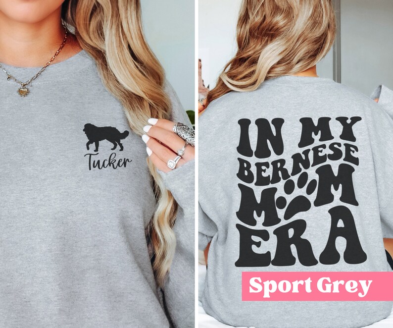 May include: A grey sweatshirt with a black silhouette of a Bernese Mountain Dog and the name "Tucker" on the front. The back of the sweatshirt has the text "In My Bernese Mom Era" in a retro font. The sweatshirt is labelled "Sport Grey".