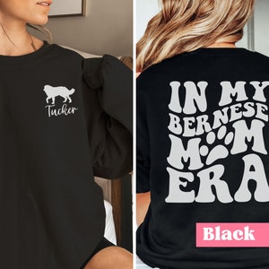 May include: Black sweatshirt with a white silhouette of a Bernese Mountain Dog and the text "Tucker" on the front. The back of the sweatshirt has the text "In My Bernese Mom Era" in a retro font with paw prints.