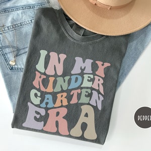 May include: A dark gray t-shirt with the text "In My Kindergarten Era" in colorful retro-style lettering.