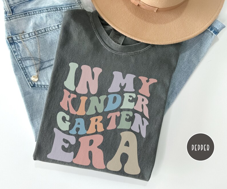 May include: A dark gray t-shirt with the text "In My Kindergarten Era" in colorful retro-style lettering.