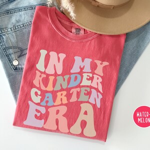 May include: A red t-shirt with the text "In My Kindergarten Era" in a retro font with rainbow colors. The shirt is laid out on a white surface with a pair of blue jeans and a brown hat.
