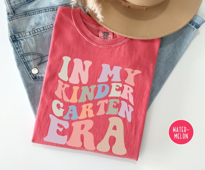 May include: A red t-shirt with the text "In My Kindergarten Era" in a retro font with rainbow colors. The shirt is laid out on a white surface with a pair of blue jeans and a brown hat.