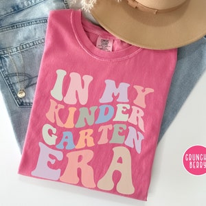 May include: Pink t-shirt with the text "In My Kinder Garden Era" in a rainbow of colors. The text is in a retro font style.