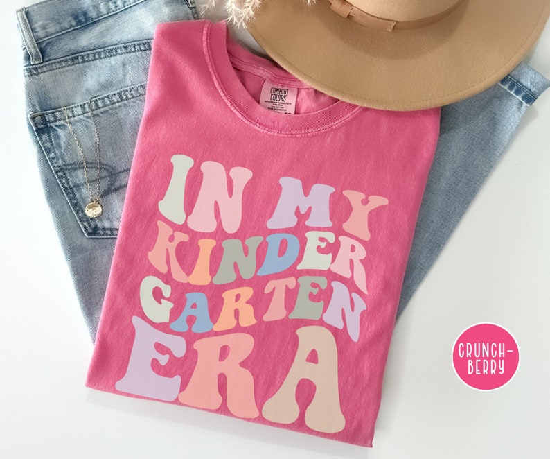 May include: Pink t-shirt with the text "In My Kinder Garden Era" in a rainbow of colors. The text is in a retro font style.