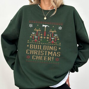 May include: A dark green sweatshirt with a Christmas-themed design featuring a pattern of hammers, nails, and stars. The text "Building Christmas Cheer!" is printed on the front of the sweatshirt.