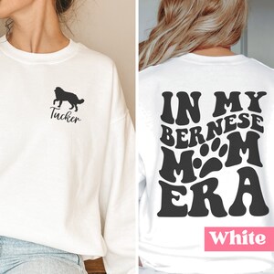 May include: White sweatshirt with a black silhouette of a dog and the name "Tucker" on the front. The back of the sweatshirt has the text "In My Bernese Mom Era" in a retro font.