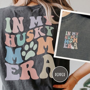 May include: A dark grey t-shirt with a colorful graphic that says "In My Husky Mom Era" with a paw print. The shirt is being worn by a person.
