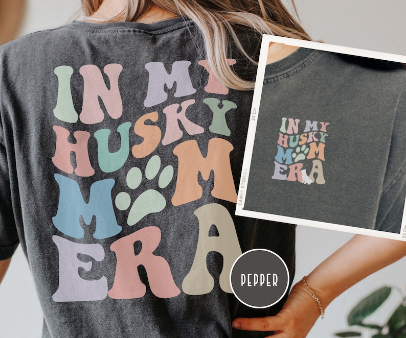 May include: A dark grey t-shirt with a colorful graphic that says "In My Husky Mom Era" with a paw print. The shirt is being worn by a person.