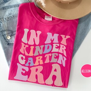 May include: Pink t-shirt with the text "In My Kindergarten Era" in rainbow colors.
