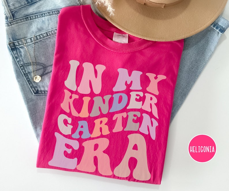 May include: Pink t-shirt with the text "In My Kindergarten Era" in rainbow colors.