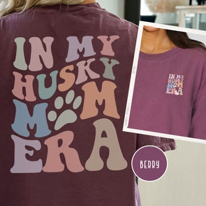 May include: A maroon long-sleeve t-shirt with the text "In My Husky Mom Era" in a retro, colorful font. The text is arranged in a circular pattern with paw prints.