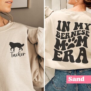 May include: A beige sweatshirt with a black silhouette of a Bernese Mountain Dog and the name "Tucker" on the front. The back of the sweatshirt has the text "In My Bernese Mom Era" in a retro font.