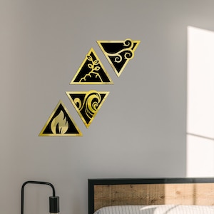 Puede incluir: Cuatro piezas de arte mural geométricas negras y doradas, cada una en forma de triángulo, que representan los elementos de fuego, tierra, agua y aire. El arte está montado en una pared gris sobre una cama con cabecero de madera y una almohada a rayas.