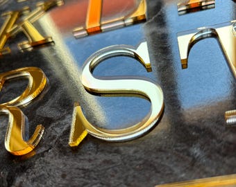 May include: Close-up of gold-colored, three-dimensional letters on a dark, textured surface. The letters appear to be reflective, with a metallic sheen. The letter "S" is in focus, with other letters like "K", "L", and "T" visible.