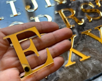 May include: Gold-coloured letter E made of acrylic. The letter is being held in a hand. The letter is part of a set of gold-coloured letters.