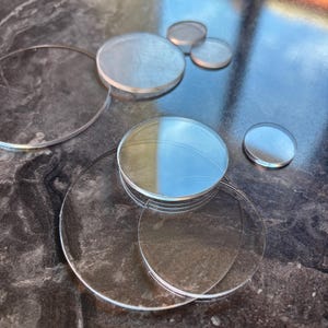 Könnte beinhalten: Eine Sammlung von klaren, kreisförmigen Acrylscheiben in verschiedenen Größen. Die transparenten, flachen, runden Teile sind in einer Gruppe angeordnet und weisen glatte Kanten sowie eine reflektierende Oberfläche auf, die das Licht einfängt.