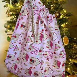 May include: A pink fabric gift bag with a gingerbread house pattern. The bag has a drawstring closure and a zipper on the side.