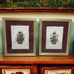 May include: Two framed floral prints with gold frames and burgundy mats. Each print features a bouquet of flowers in a terracotta pot, with a white background and a handwritten signature.