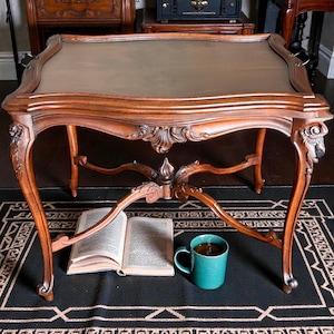 May include: An antique wooden coffee table with a brown leather top. The table has ornate carvings and curved legs. A teal mug of tea and an open book rest on a black and white patterned rug beneath the table.