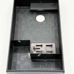 May include: A rectangular black plastic storage tray featuring a textured geometric pattern on the base. The interior includes a raised rectangular compartment with four vertical prongs, a circular cutout near the top edge, and a raised shelf section.