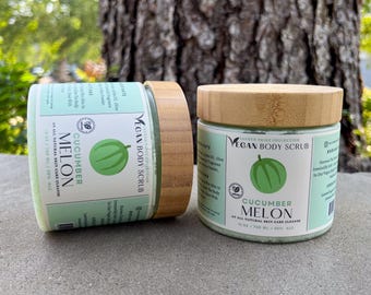 May include: Two jars of cucumber melon vegan body scrub with wooden lids. The labels are light green with the text "Cucumber Melon" and a watermelon graphic. The jars are 100 ml / 3.8% ALC.