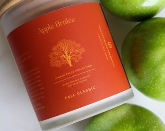 Puede incluir: Un tarro de vela rojo con una tapa blanca y una parte superior de madera. El tarro tiene una etiqueta que dice "Apple Brûlée" y "Lauren Paige Collection". La vela está rodeada de manzanas verdes.