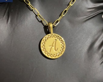 Puede incluir: Collar dorado con un colgante circular con la letra "A" en escritura cursiva. El colgante está rodeado por un borde trenzado y cuelga de una cadena con eslabones rectangulares. El collar se muestra sobre un fondo oscuro.